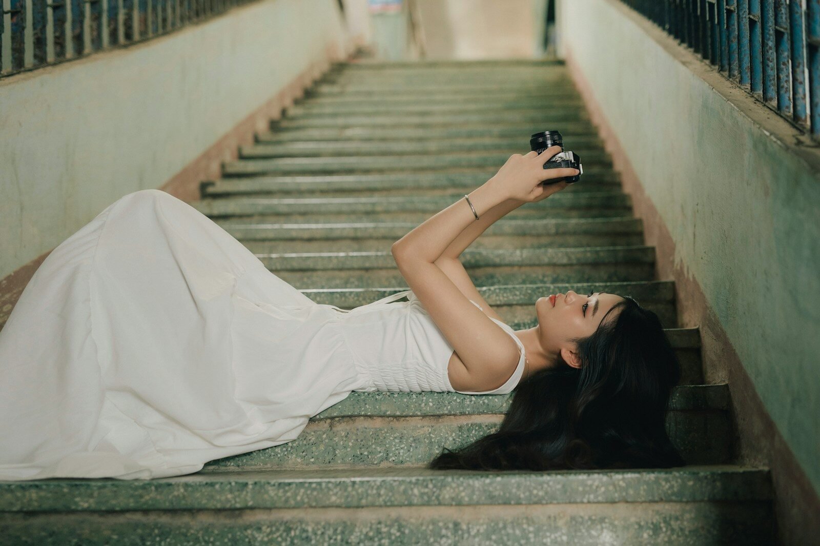 A woman in a white dress lying on concrete stairs holding a camera